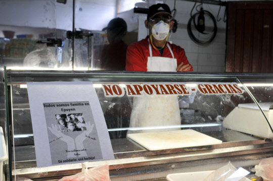 A man wearing a mask waits for clients at his shop, with a sign posted that reads in Spanish "We are all family in Epuyen, be supportive, use a mask" in Epuyen, Argentina, Friday, Jan. 11, 2019. An Argentine judge has ordered 85 residents of a remote Patagonian town to stay in their homes for at least 30 days to help halt an outbreak of hantavirus in which nine people have died. (AP Photo/Gustavo Zaninelli)